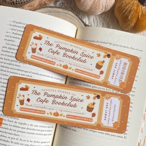 Könnte beinhalten: Zwei orangefarbene und cremefarbene Lesezeichen mit dem Text "The Pumpkin Spice Cafe Bookclub" und "Admit One cozy reading night". Die Lesezeichen liegen auf einem offenen Buch, mit dekorativen Kürbissen und einer Kerze.