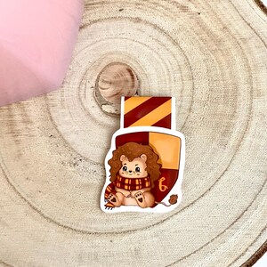 May include: A Gryffindor house bookmark featuring a cartoon lion wearing a red and gold scarf. The bookmark is shaped like a shield with the Gryffindor house crest.