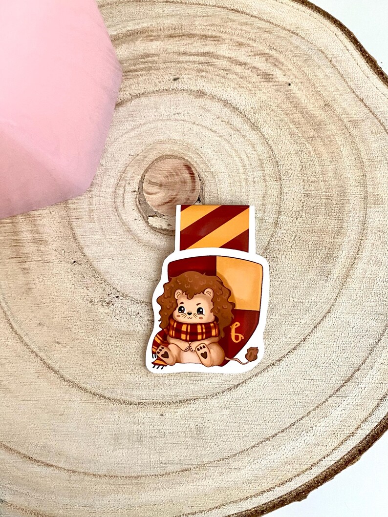 May include: A Gryffindor house bookmark featuring a cartoon lion wearing a red and gold scarf. The bookmark is shaped like a shield with the Gryffindor house crest.