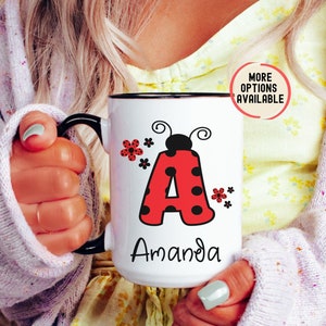 May include: White ceramic mug with a black handle and rim. The mug features a red letter "A" with black spots, designed to resemble a ladybug, along with the name "Amanda" in black script. Red flowers and the text "MORE OPTIONS AVAILABLE" are also visible.