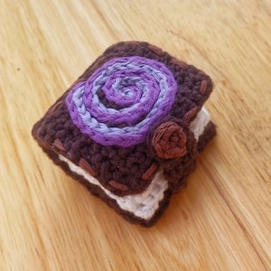 Puede incluir: Un pequeño libro de ganchillo con un diseño de espiral morado y azul en la portada. El libro es marrón y blanco y tiene una pequeña flor de ganchillo marrón en el lateral.