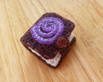 Op de afbeelding: Een klein gehaakt boekje met een paars en blauw swirl-ontwerp op de omslag. Het boekje is bruin en wit en heeft een klein bruin gehaakt bloemetje aan de zijkant.