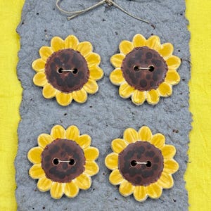 Puede incluir: Cuatro botones con forma de girasol, con pétalos amarillos y centros marrones, están dispuestos sobre un papel gris texturizado. Los botones tienen dos agujeros para coser. Un lazo de cuerda está atado en la parte superior. El fondo es amarillo.