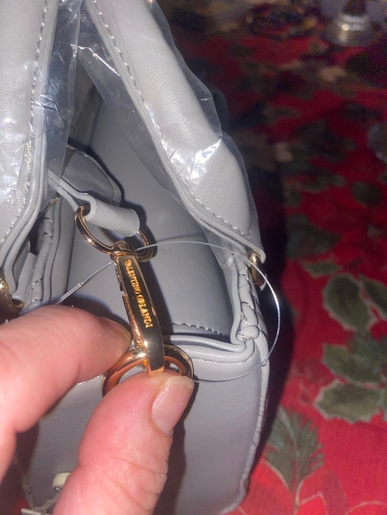 May include: A light gray handbag with gold-colored hardware. The bag is made of a smooth material and features a top handle and a detachable shoulder strap. The brand name "Valentino Orlandi" is visible on the clasp.