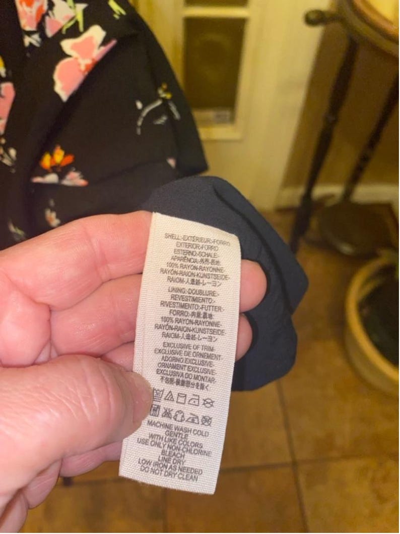 May include: A black and white care label with instructions for washing and caring for a garment. The label says "Machine wash cold with like colours, gentle, use only non-chlorine bleach, low iron as needed, line dry, do not dry clean."