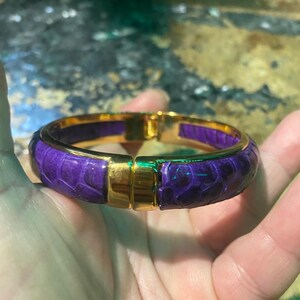 May include: A gold-toned hinged bangle bracelet with a purple, textured, snake-skin pattern. The bracelet has a rectangular clasp and a gold-toned interior. The bracelet is a fashion accessory.