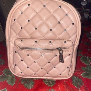 Op de afbeelding: Een blush-kleurige rugzak met een gewatteerd patroon en zilverkleurige stud accenten. De kleine voorzak heeft een ritssluiting. De tas heeft een handvat aan de bovenkant en verstelbare bandjes. De tas staat op een rode bloemen tafelkleed.