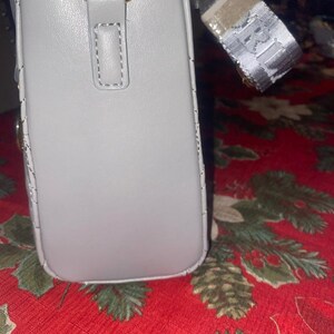 May include: A light gray rectangular handbag with a gold-colored metal clasp and a patterned strap. The bag has a stitched design along the edges and a small loop on the front. The strap has a repeating pattern.