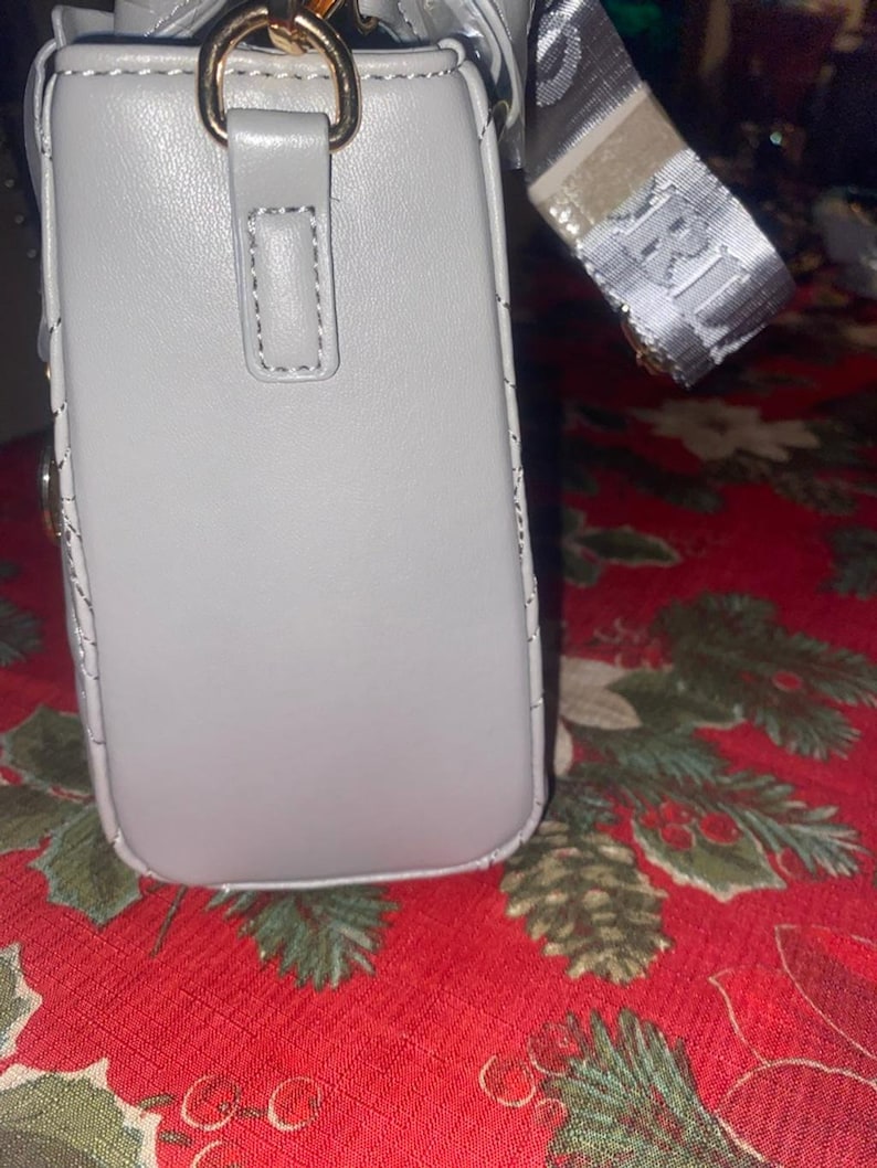 May include: A light gray rectangular handbag with a gold-colored metal clasp and a patterned strap. The bag has a stitched design along the edges and a small loop on the front. The strap has a repeating pattern.
