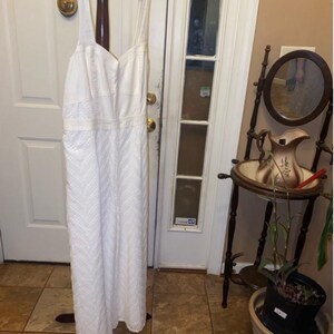 May include: A white maxi dress with a delicate embroidered pattern. The dress has a sweetheart neckline and thin straps. It is hanging on a wooden hanger.