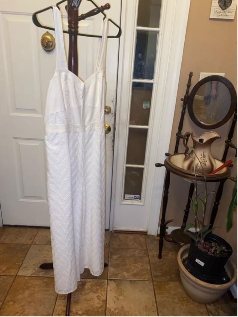 May include: A white maxi dress with a delicate embroidered pattern. The dress has a sweetheart neckline and thin straps. It is hanging on a wooden hanger.