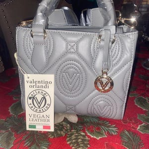 May include: A gray, quilted vegan leather handbag with a top handle and gold-colored hardware. The bag features the Valentino Orlandi logo and a charm. The tag reads "VEGAN LEATHER DESIGNED IN ITALY."