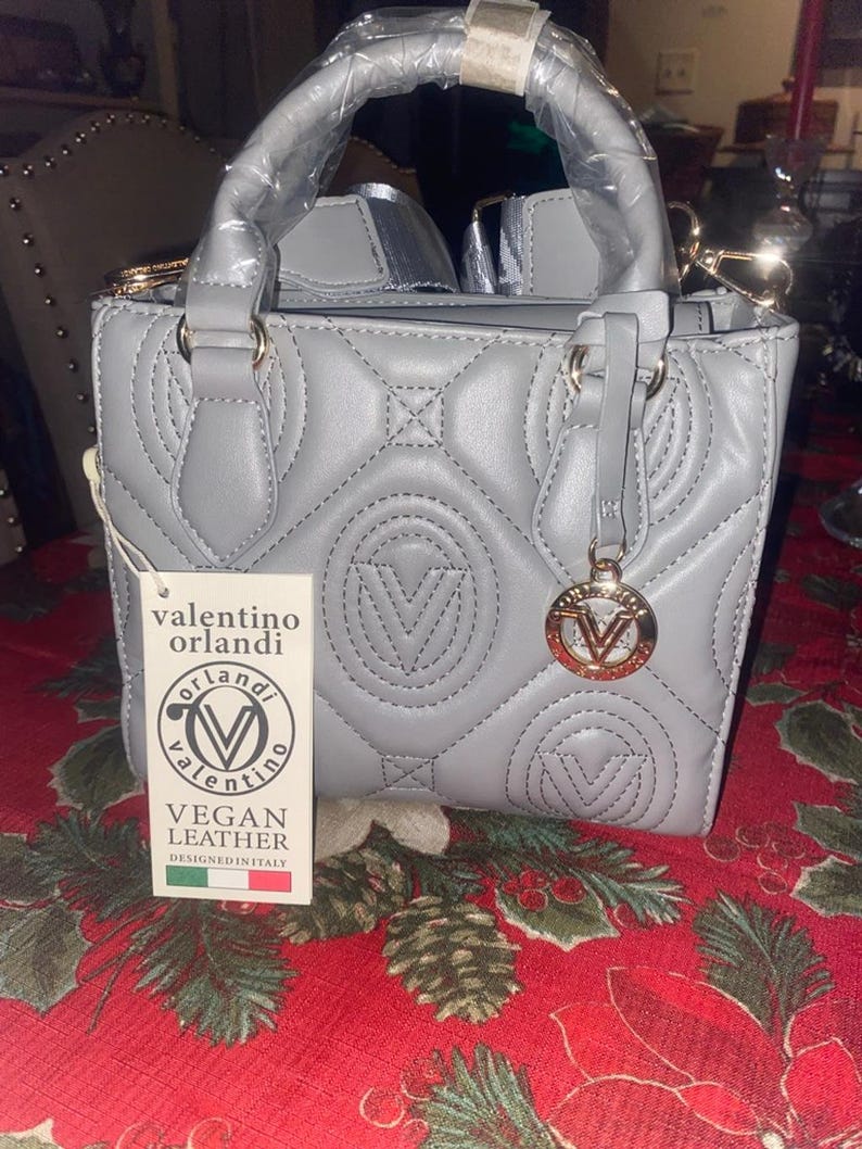 May include: A gray, quilted vegan leather handbag with a top handle and gold-colored hardware. The bag features the Valentino Orlandi logo and a charm. The tag reads "VEGAN LEATHER DESIGNED IN ITALY."