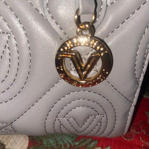 May include: Close-up of a gray quilted handbag with a gold-colored circular charm. The charm features a stylized letter "V" in the center. The stitching on the bag is silver.