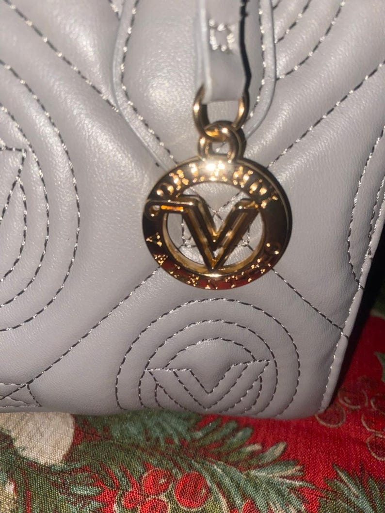 May include: Close-up of a gray quilted handbag with a gold-colored circular charm. The charm features a stylized letter "V" in the center. The stitching on the bag is silver.