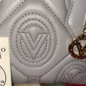May include: Close-up of a gray quilted handbag with black stitching. The bag features a prominent stitched "V" logo and a gold-colored charm with the brand's logo. The bag's design includes a repeating pattern of stitched shapes.