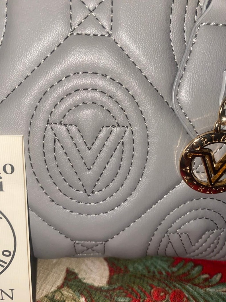 May include: Close-up of a gray quilted handbag with black stitching. The bag features a prominent stitched "V" logo and a gold-colored charm with the brand's logo. The bag's design includes a repeating pattern of stitched shapes.