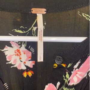 May include: Black floral dress with a button fastening and a Free People label. The dress features a large floral print with pink, white and green flowers on a black background. The label reads "Free People" and "Made in Indonesia".