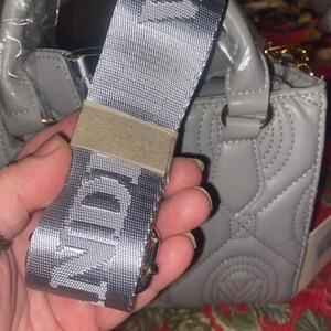 May include: A gray handbag with a matching shoulder strap. The strap is silver with the letters "NDI" printed on it. The handbag has a quilted design and gold-colored hardware.