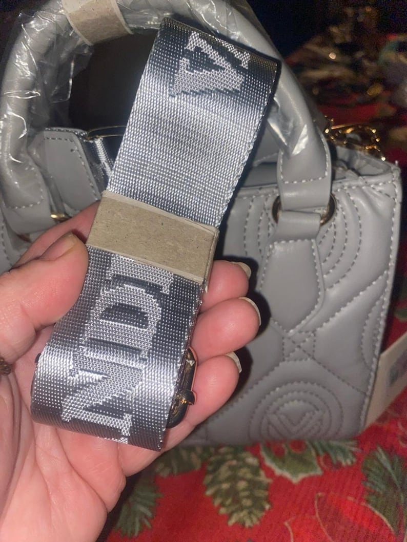 May include: A gray handbag with a matching shoulder strap. The strap is silver with the letters "NDI" printed on it. The handbag has a quilted design and gold-colored hardware.
