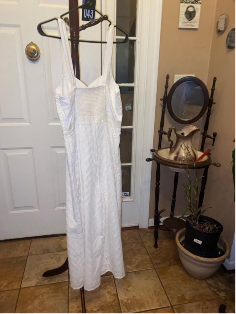 May include: White eyelet maxi dress with a sweetheart neckline and spaghetti straps. The dress is hanging on a wooden hanger.