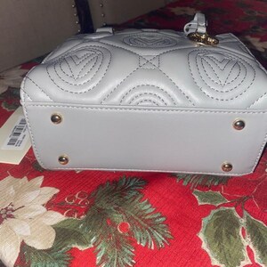 May include: A light gray, quilted handbag with a top handle and gold-colored hardware. The bag features a repeating heart-shaped pattern stitched into the leather. The base of the bag has four gold-colored feet.