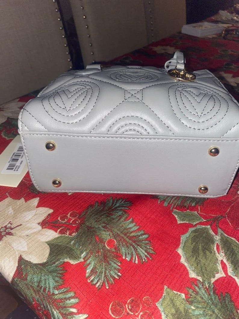 May include: A light gray, quilted handbag with a top handle and gold-colored hardware. The bag features a repeating heart-shaped pattern stitched into the leather. The base of the bag has four gold-colored feet.