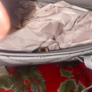 May include: Close-up of a light blue quilted handbag with a gold-colored clasp. The interior is filled with crumpled white tissue paper. The bag is on a red surface with green floral patterns.