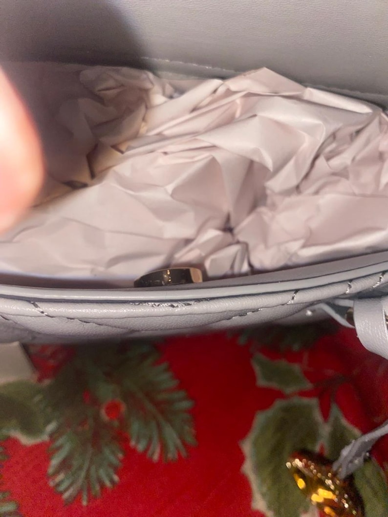 May include: Close-up of a light blue quilted handbag with a gold-colored clasp. The interior is filled with crumpled white tissue paper. The bag is on a red surface with green floral patterns.