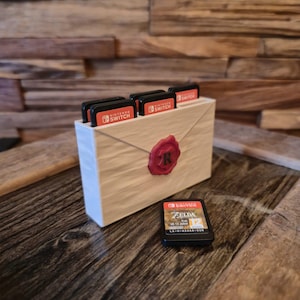 Op de afbeelding: Een houten Nintendo Switch game cartridge houder in de vorm van een envelop, met een rode zegel. De houder bevat meerdere game cartridges. Eén game cartridge is buiten de houder geplaatst. De houder staat op een houten oppervlak.