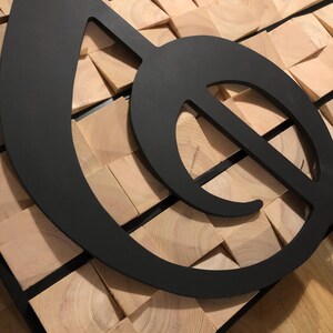 Op de afbeelding: Een grote, zwarte metalen G-sleutel, een muzieksymbool, gemonteerd op een moza&iuml;ekachtergrond van lichtbruine houten blokken. De G-sleutel is een gedurfd, modern ontwerp en het hout biedt een natuurlijke, getextureerde achtergrond.