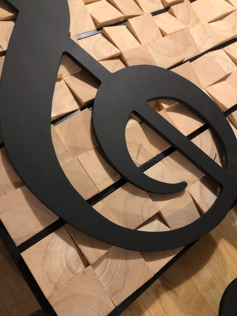 Op de afbeelding: Een grote, zwarte metalen G-sleutel, een muzieksymbool, gemonteerd op een moza&iuml;ekachtergrond van lichtbruine houten blokken. De G-sleutel is een gedurfd, modern ontwerp en het hout biedt een natuurlijke, getextureerde achtergrond.