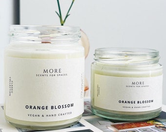 Peut inclure: Deux pots de bougies en verre avec des étiquettes blanches. Les étiquettes indiquent "MORE SCENTS FOR SPACES" et "ORANGE BLOSSOM VEGAN & HAND CRAFTED".