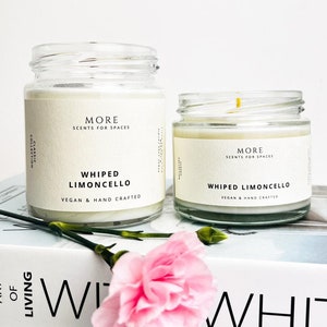 Peut inclure: Deux bougies en pot de verre avec l'inscription "WHIPPED LIMONCELLO" et "VEGAN & HAND CRAFTED" sur l'étiquette. Les bougies sont posées sur un livre avec le texte "ART OF LIVING" et une fleur rose.