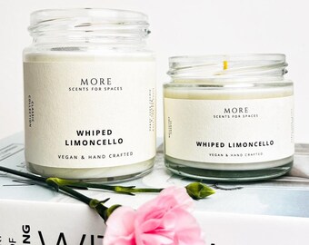 May include: Two glass jar candles with the text "WHIPPED LIMONCELLO" and "VEGAN & HAND CRAFTED" on the label. The candles are on a book with the text "ART OF LIVING" and a pink flower.