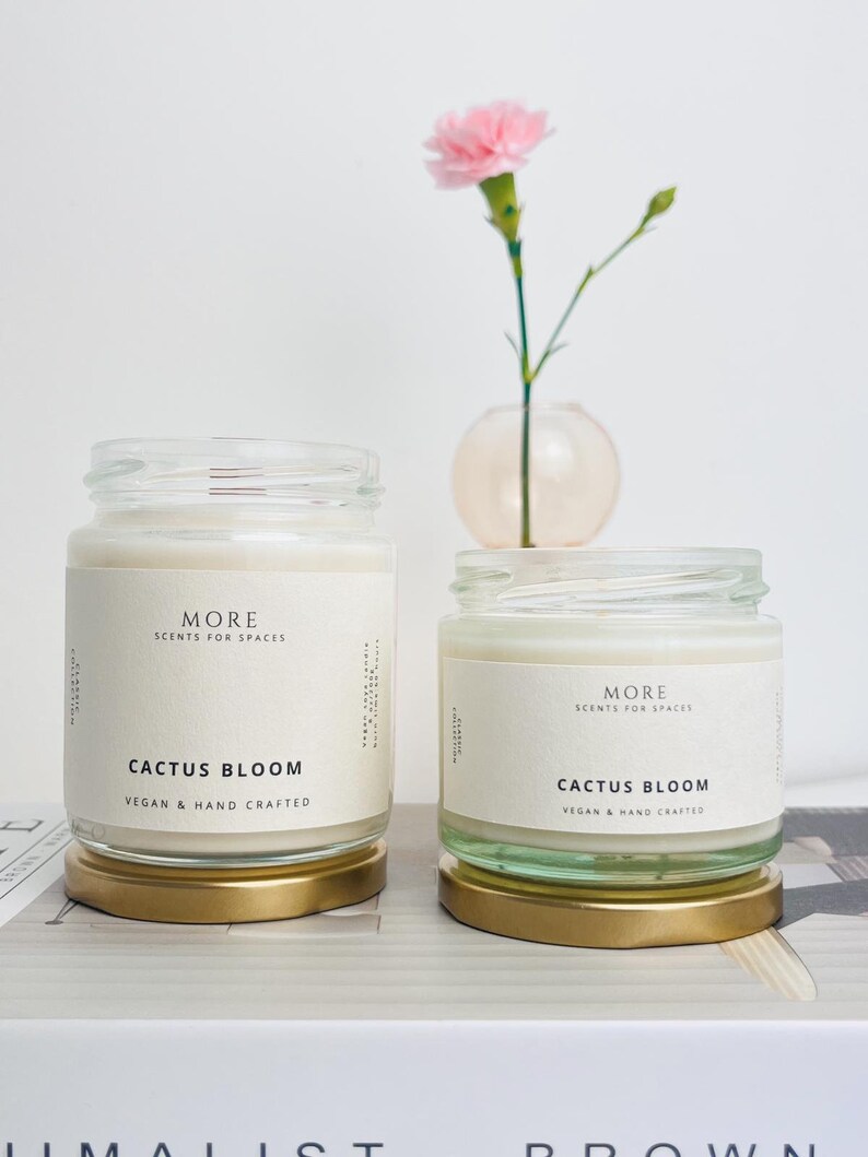 May include: Two glass candle jars with gold lids. The jars are filled with white wax and have labels that read "MORE SCENTS FOR SPACES" and "CACTUS BLOOM VEGAN & HAND CRAFTED".