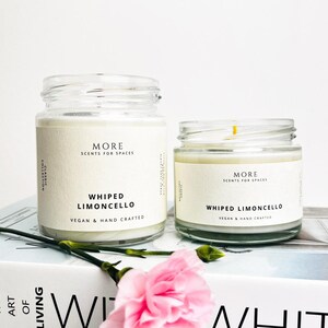 May include: Two glass candle jars with white labels. The labels read "MORE SCENTS FOR SPACES" and "WHIPPED LIMONCELLO VEGAN & HAND CRAFTED". The candles are sitting on a white book with the word "WHIT" visible.