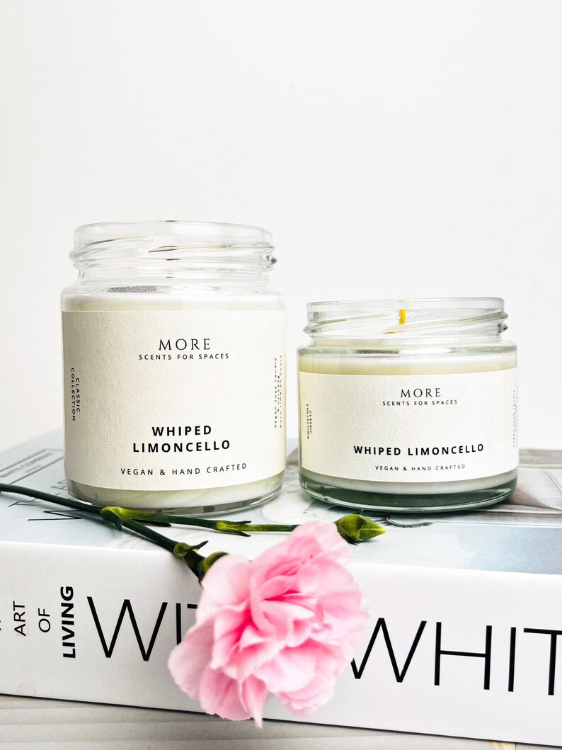 May include: Two glass candle jars with white labels. The labels read "MORE SCENTS FOR SPACES" and "WHIPPED LIMONCELLO VEGAN & HAND CRAFTED". The candles are sitting on a white book with the word "WHIT" visible.