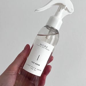 Peut inclure: Une bouteille en verre transparente avec une buse de pulv&eacute;risation blanche remplie d'un liquide rose clair. La bouteille porte une &eacute;tiquette blanche avec le texte "MORE SCENTS FOR SPACES" et "TEA ROSE Linen Room Spray 150 ml".
