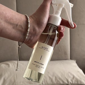 Peut inclure: Une bouteille en verre transparente avec une buse de pulv&eacute;risation blanche et une &eacute;tiquette blanche. L'&eacute;tiquette indique "MORE SCENTS FOR SPACES" et "WHITE LINEN Linen Room Spray 150 ml".