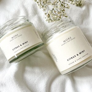May include: Two glass candle jars with white labels. The labels read "MORE SCENTS FOR SPACES" and "CITRUS & MINT" with the text "VEGAN & HAND CRAFTED" below. The jars are sitting on a white fabric surface.