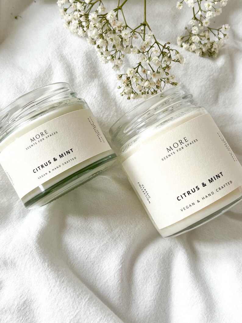 May include: Two glass candle jars with white labels. The labels read "MORE SCENTS FOR SPACES" and "CITRUS & MINT" with the text "VEGAN & HAND CRAFTED" below. The jars are sitting on a white fabric surface.