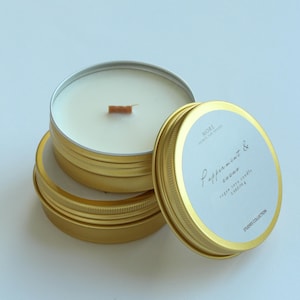 Peut inclure: Boîtes de bougies dorées avec des étiquettes blanches et du texte. Les boîtes sont empilées, avec un couvercle partiellement ouvert, révélant une bougie blanche et une mèche en bois. L'étiquette indique "Peppermint & cacao" et "vegan soya candle".