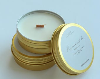Peut inclure: Boîtes de bougies dorées avec des étiquettes blanches et du texte. Les boîtes sont empilées, avec un couvercle partiellement ouvert, révélant une bougie blanche et une mèche en bois. L'étiquette indique "Peppermint & cacao" et "vegan soya candle".