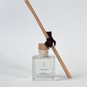 Op de afbeelding: Een heldere glazen rietverspreiderfles met een houten dop en houten rietjes. De fles heeft een label met "Island Hibiscus" en "MORE SCENTS FOR SPACES". Een donkerrood lint is om de rietjes gebonden. Voor het parfumeren van ruimtes.