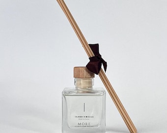 May include: A clear glass reed diffuser bottle with a wooden cap and a bundle of wooden reeds. The bottle has a label that reads "Island Hibiscus" and "MORE SCENTS FOR SPACES". A dark red ribbon is tied around the reeds.