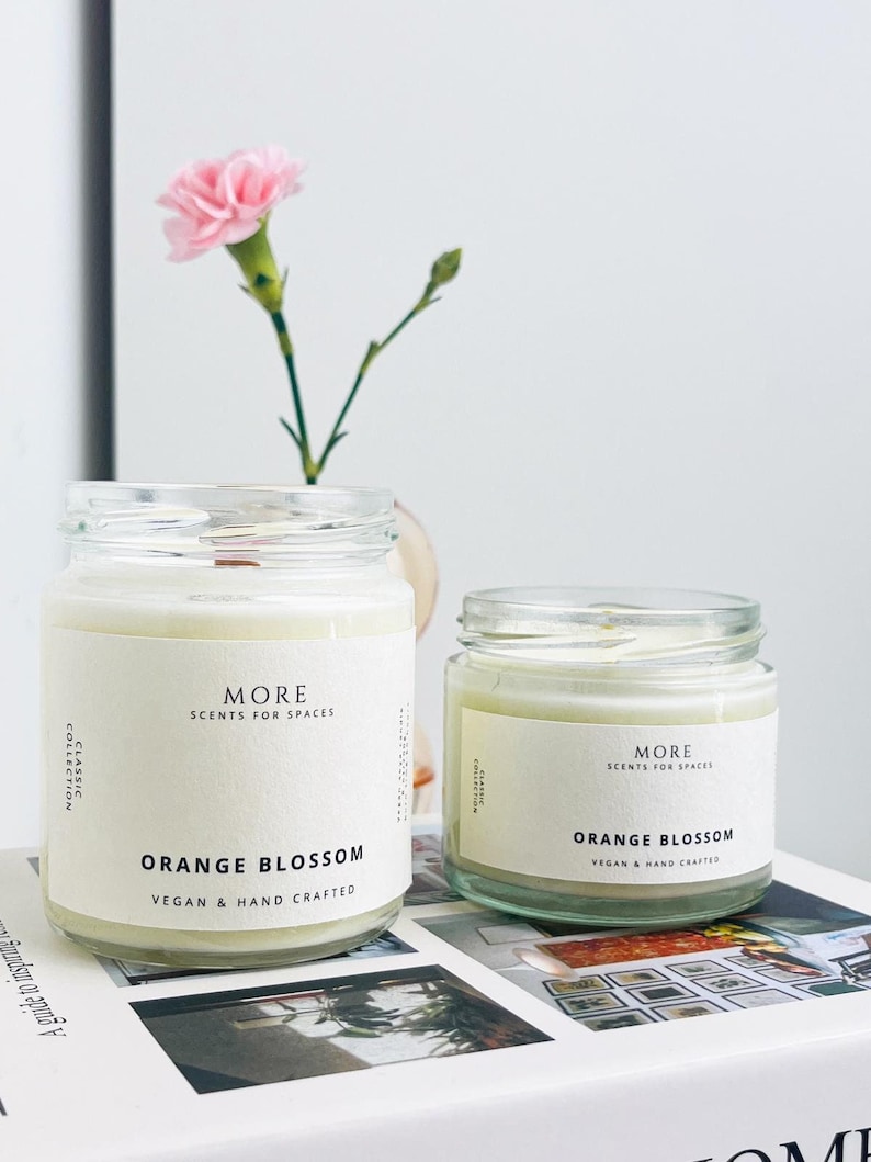 May include: Two glass candle jars with white labels. The labels read "MORE SCENTS FOR SPACES" and "ORANGE BLOSSOM VEGAN & HAND CRAFTED".