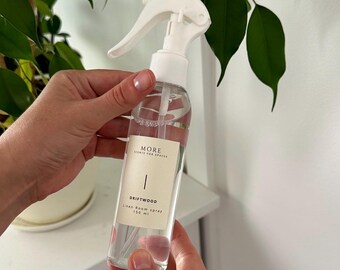 May include: A clear glass bottle with a white spray nozzle and a white label. The label says "MORE SCENTS FOR SPACES" and "DRIFTWOOD Linen Room Spray 150 ml".