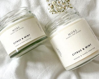 Peut inclure: Deux bougies en pot de verre avec de la cire blanche et des étiquettes indiquant "MORE SCENTS FOR SPACES CITRUS & MINT VEGAN & HAND CRAFTED". Les bougies sont sur un tissu blanc, avec des fleurs blanches en arrière-plan.