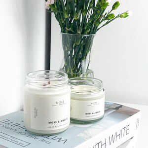 May include: Two white candle jars with black labels that read "More Scents for Spaces" and "Moss & Amber" on a stack of books with the text "The Art of Living With White" on the top book.